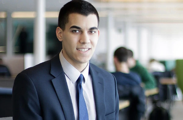 A young man in a suit smiling at the camera with an office environment in the background.